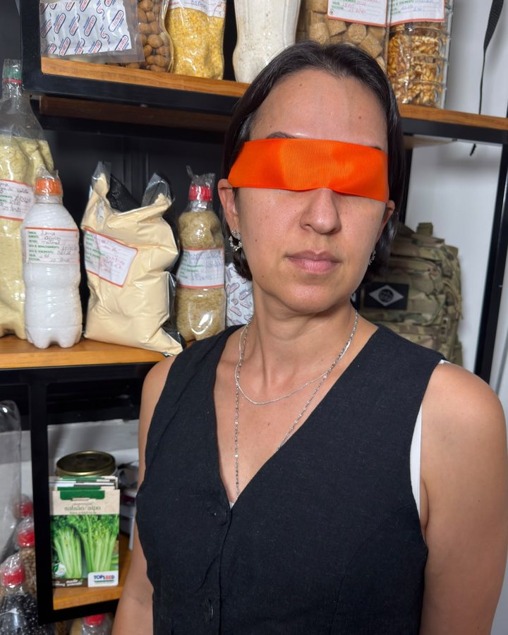 Cinthia de venda nos olhos, em frente à despensa cheia de garrafas PET e mantimentos armazenados.